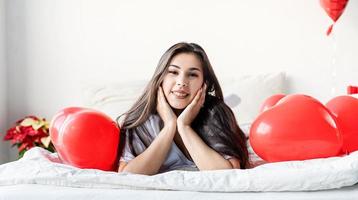 Joven mujer morena feliz acostado en la cama con globos en forma de corazón rojo foto