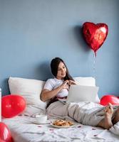 Joven mujer morena feliz sentada en la cama con globos en forma de corazón rojo charlando con su novio en la computadora portátil que muestra el gesto del corazón con las manos foto