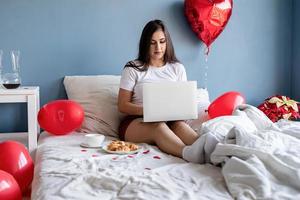 Feliz joven mujer morena sentada en la cama con globos en forma de corazón rojo trabajando en la computadora portátil foto