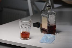 vaso con alcohol, botella de whisky y mascarilla médica sobre la mesa. concepto de eventos sociales cancelados durante la cuarentena por el virus de la corona y la pandemia