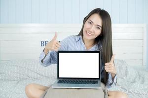 Joven mujer asiática sonriendo y mostrando la pantalla de la computadora portátil en blanco en su dormitorio foto