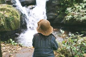 chica hipster con estilo viajando en el bosque, vista posterior. mujer joven con sombrero explorando en el bosque de verano, mirando al río. concepto de viaje y pasión por los viajes foto