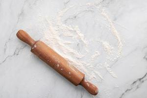 Scattered wheat flour and rolling pin set up on white marble background flat lay.