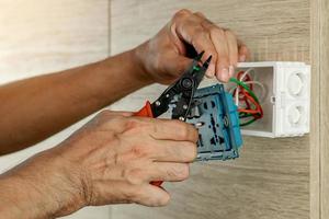 Electrician is stripping electrical wires in a plastic box on a wooden wall to install the electrical outlet. photo