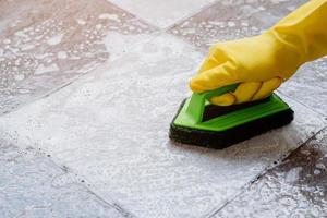 Human hands wearing yellow rubber gloves are using a green color plastic floor scrubber to scrub the tile floor with a floor cleaner. photo