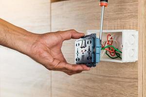 Hand of an electrician is using a screwdriver to attach the wires to the universal wall outlet AC power plug with USB port and on-off in a plastic box on a wooden wall. photo