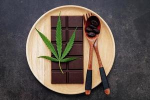 Imagen conceptual de alimentos de hoja de cannabis con chocolate negro y tenedor sobre fondo de hormigón oscuro. foto