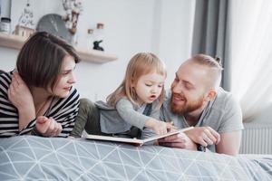 Father, Mother and Little Daughter Reading Children's Book on a Sofa in the Living Room. Happy big family read an interesting book on a festive day. Parents love their children photo