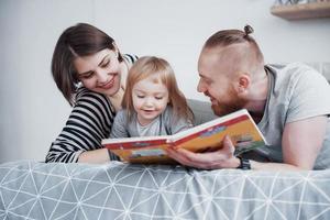 padre, madre e hija leyendo un libro para niños en un sofá en la sala de estar. gran familia feliz lee un libro interesante en un día festivo. los padres aman a sus hijos foto