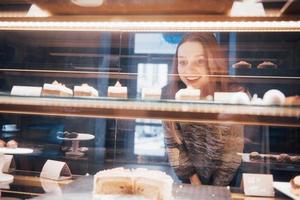 Mujer sonriente a la cámara a través de la vitrina con dulces y tortas en el interior de la cafetería moderna