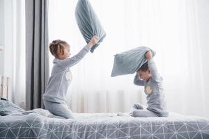 niño y niña organizaron una pelea de almohadas en la cama del dormitorio. los niños traviesos se golpean las almohadas. les gusta ese tipo de juego foto