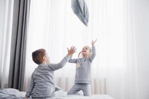 Kids playing in parents bed. Children wake up in sunny white bedroom. Boy and girl play in matching pajamas. Sleepwear and bedding for child and baby. Nursery interior for toddler kid. Family morning photo