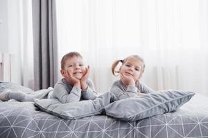 Happy kids playing in white bedroom. Little boy and girl, brother and sister play on the bed wearing pajamas. Nursery interior for children. Nightwear and bedding for baby and toddler. Family at home photo