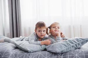 Happy kids playing in white bedroom. Little boy and girl, brother and sister play on the bed wearing pajamas. Nursery interior for children. Nightwear and bedding for baby and toddler. Family at home photo