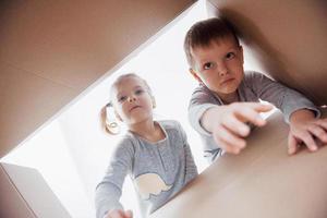 Dos niños pequeños, niño y niña, abriendo una caja de cartón y mirando hacia adentro con sorpresa. foto