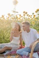 Young couple having picnic on sunflower field at sunset