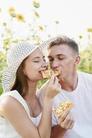 Young couple having picnic on sunflower field at sunset, biting a piece of pizza together photo