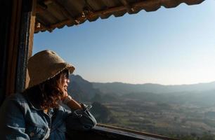Young asian woman sitting in the hotel room near window, looking at the view of moutain and drink coffee in morning. tourism and vacation concept photo
