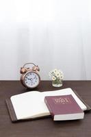 Bible and notebook on the table with an alarm clock by the window. Vertical format photo