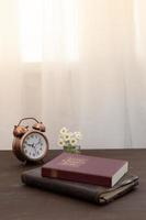 Bible and notebook on the table with an alarm clock by the window. Vertical format photo