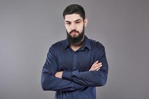 Young handsome man leaning against grey wall with arms crossed. A serious young man with a beard looks at the camera photo