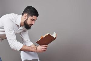 Portrait of a bearded young man wearing a white shirt and holding an open planner and a pen. A gray wall background photo