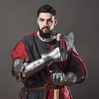 Medieval knight on grey background. Portrait of brutal dirty face warrior with chain mail armour red and black clothes and battle axe photo