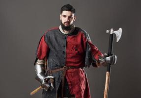 Medieval knight on grey background. Portrait of brutal dirty face warrior with chain mail armour red and black clothes and battle axe photo