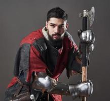 Medieval knight on grey background. Portrait of brutal dirty face warrior with chain mail armour red and black clothes and battle axe photo