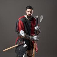 Medieval knight on grey background. Portrait of brutal dirty face warrior with chain mail armour red and black clothes and battle axe photo