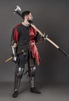 Medieval knight on grey background. Portrait of brutal dirty face warrior with chain mail armour red and black clothes and battle axe photo