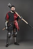 Medieval knight on grey background. Portrait of brutal dirty face warrior with chain mail armour red and black clothes and battle axe photo