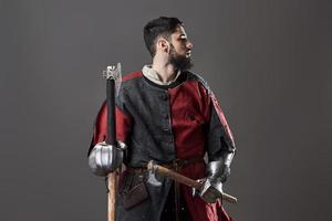 Medieval knight on grey background. Portrait of brutal dirty face warrior with chain mail armour red and black clothes and battle axe photo