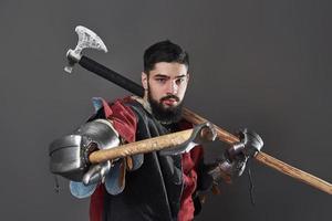 Medieval knight on grey background. Portrait of brutal dirty face warrior with chain mail armour red and black clothes and battle axe photo