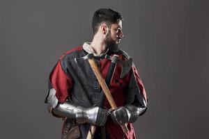 Medieval knight on grey background. Portrait of brutal dirty face warrior with chain mail armour red and black clothes and battle axe photo