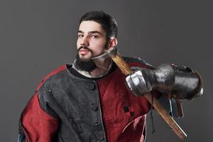 Medieval knight on grey background. Portrait of brutal dirty face warrior with chain mail armour red and black clothes and battle axe photo