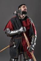 Medieval knight on grey background. Portrait of brutal dirty face warrior with chain mail armour red and black clothes and battle axe photo