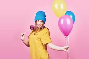 Cute funny girl in blue cap portrait holds an air colorful balloons and lollipop smiling on pink background. Beautiful multicultural Caucasian girl smiling happy photo