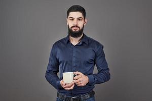 Hombre guapo con barba con barba y bigote de pelo elegante en la cara seria en camisa sosteniendo una taza blanca o una taza bebiendo té o café en el estudio sobre fondo gris foto