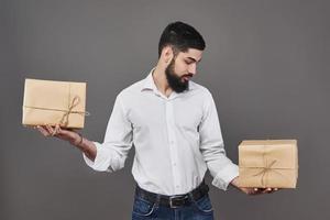 guapo chico romántico está mirando la caja y hace una elección. sosteniendo una caja de regalo de dos grandes para su pareja, sobre fondo gris.