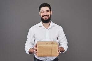 With love for you. Good looking young man in blue jeans shirt holding a gift box and looking at camera while standing against grey background photo