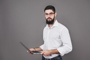 Handsome business man in glasses and suit holding laptop in hands and writing something. Side view. Isolated gray background photo