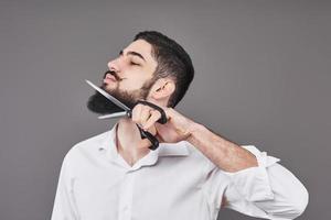 No more beard. Portrait of handsome young man cutting his beard with scissors and looking at camera while standing against grey background. New trend photo