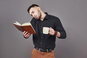 Portrait of young handsome man with phone, cup of coffe and book on hands. The concept of overtime, stress and a lot of work photo