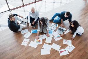 Planning strategy together. Business team looking at papers on floor with manager pointing to one idea. Cooperation corporate achievement. Planning design draw. Teamwork concept photo