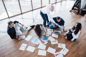 Planning strategy together. Business team looking at papers on floor with manager pointing to one idea. Cooperation corporate achievement. Planning design draw. Teamwork concept photo