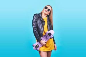 Attractive tanned girl with smile in face wears black leather jacket posing with pleasure. Indoor photo of lovely woman in sunglasses standing with skateboard on blue background