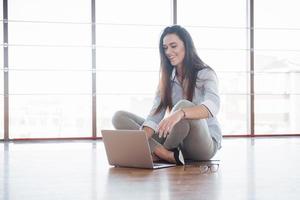 Vista lateral de una chica atractiva usando una computadora portátil en el área wifi pública y sonriendo mientras está sentada en el piso
