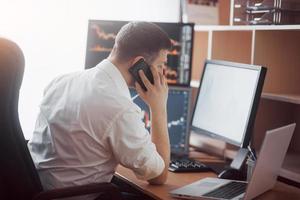 Over the shoulder view of and stock broker trading online while accepting orders by phone. Multiple computer screens ful of charts and data analyses in background photo