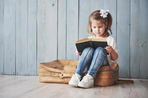 listo para grandes viajes. niña feliz leyendo un libro interesante con un maletín grande y sonriendo. concepto de viaje, libertad e imaginación. foto